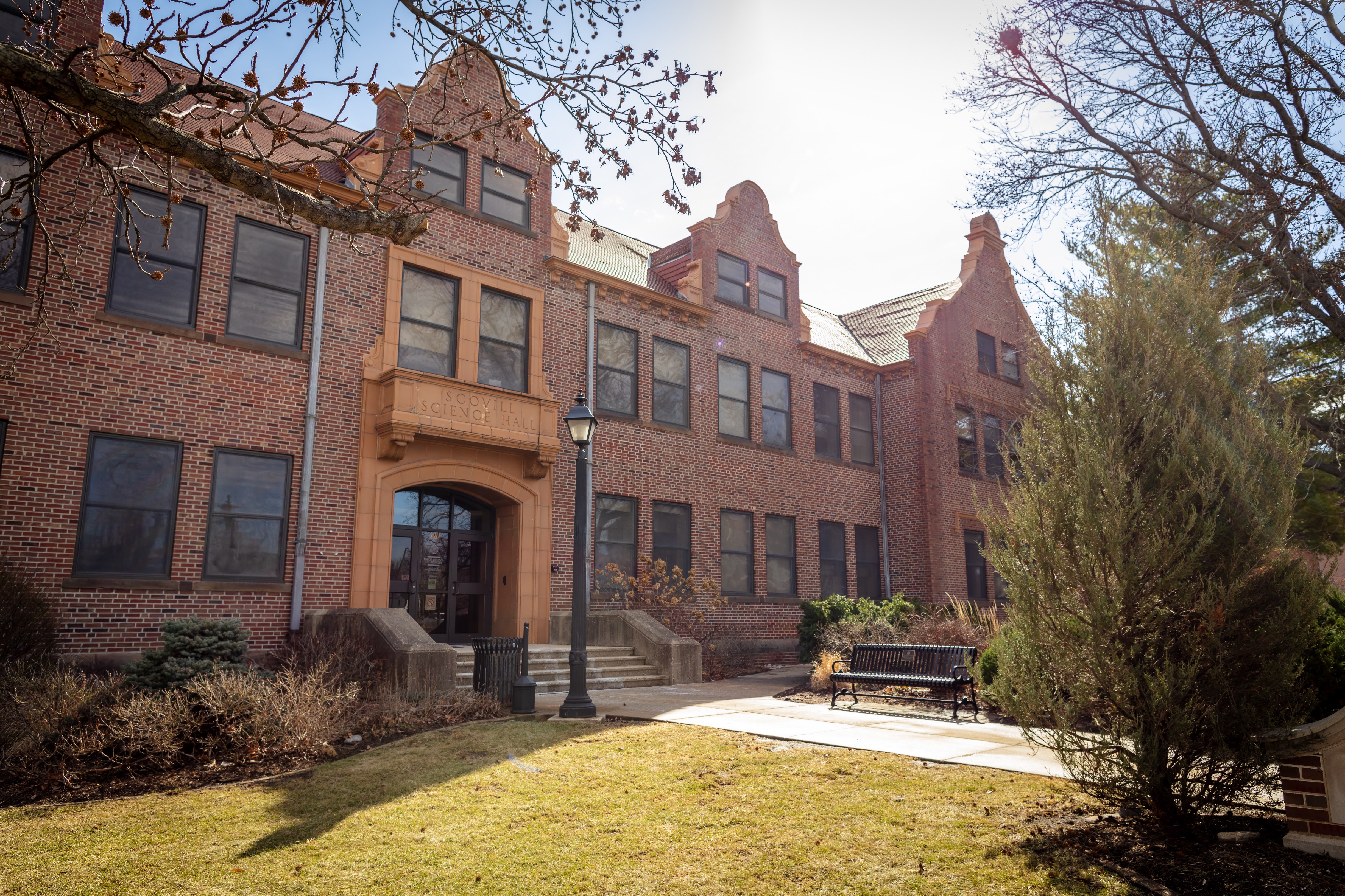 Exterior view of Scovill Science Hall, a historic three-story red-brick academic building with decorative gables and stone trim around the main entrance. A walkway leads to the front doors, with a lamp post, bench, grass lawn, and landscaped shrubs in the foreground. Bare tree branches frame the scene, and bright sunlight illuminates the building on a clear day. Visible text above the entrance reads “Scovill Science Hall.”