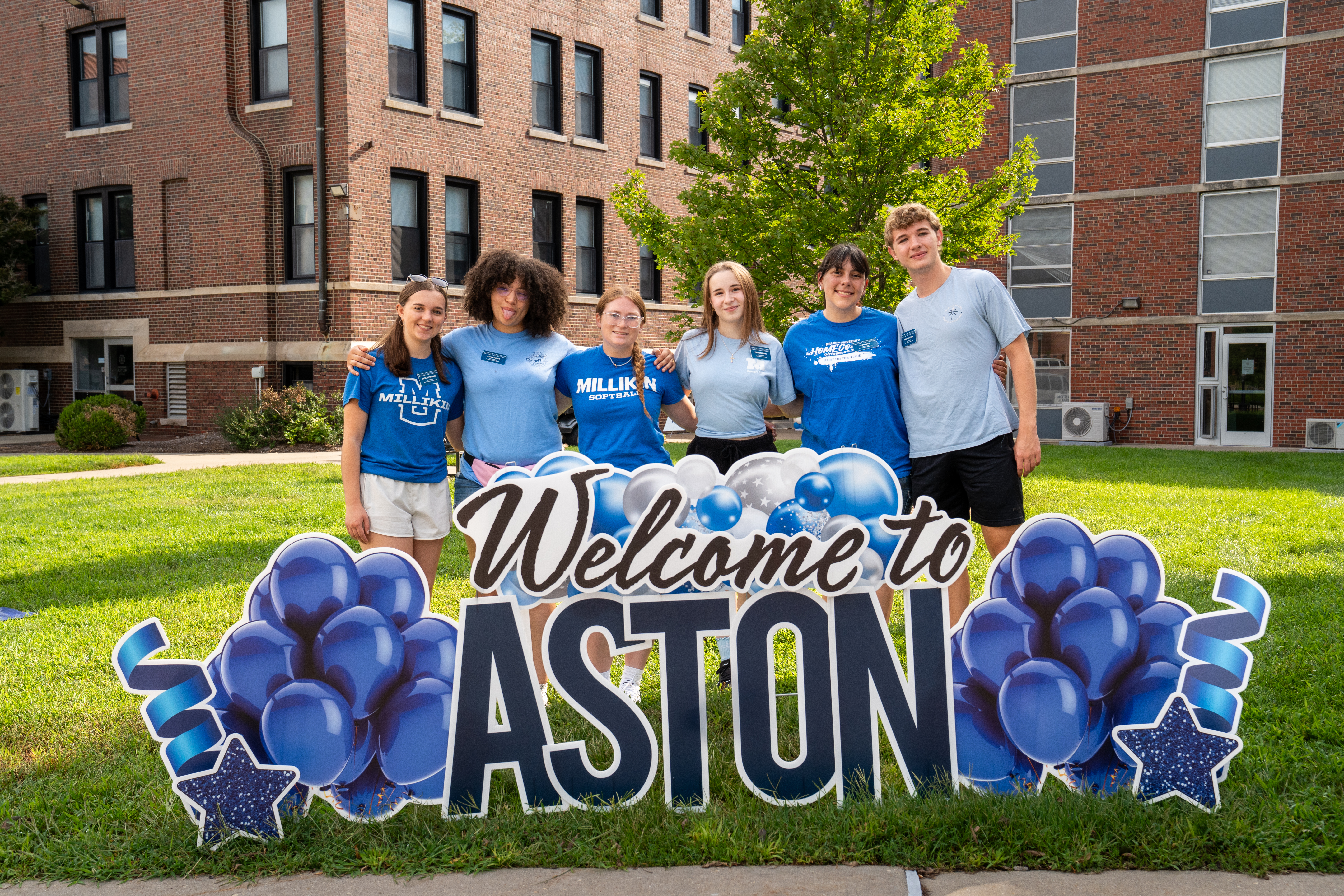 Six Millikin University students stand on a lawn in front of a residence hall, posing behind a large “Welcome to ASTON” sign decorated with blue balloons, stars, and streamers.