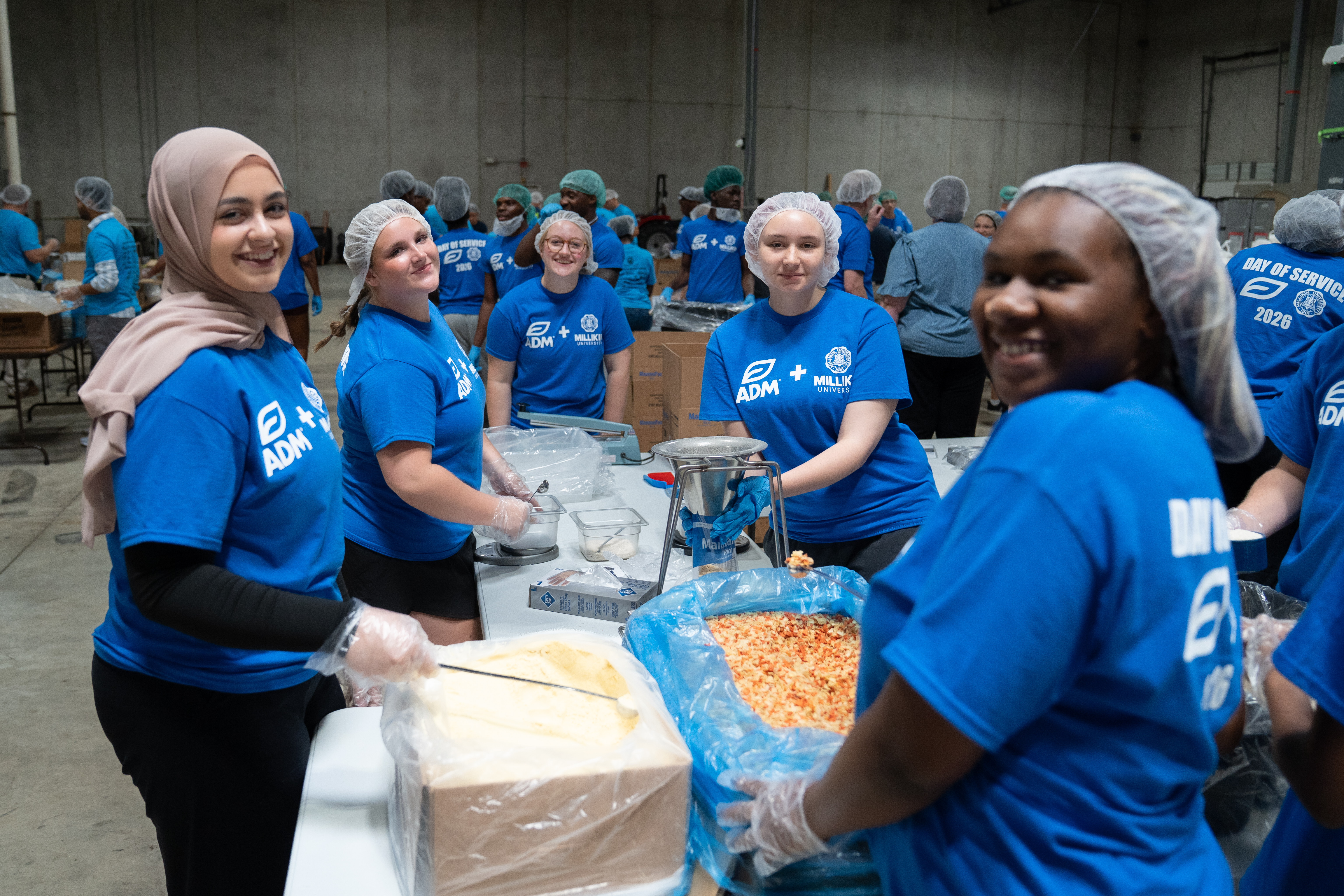 Volunteers in matching blue ADM-Millikin Day of Service T-shirts and hairnets work together at a food-packing station inside a warehouse. Several volunteers stand around a table with large containers of ingredients, scales, and packaging supplies, while additional volunteers work at stations throughout the background. The shirts display the text “ADM + Millikin University” and “Day of Service 2026.”