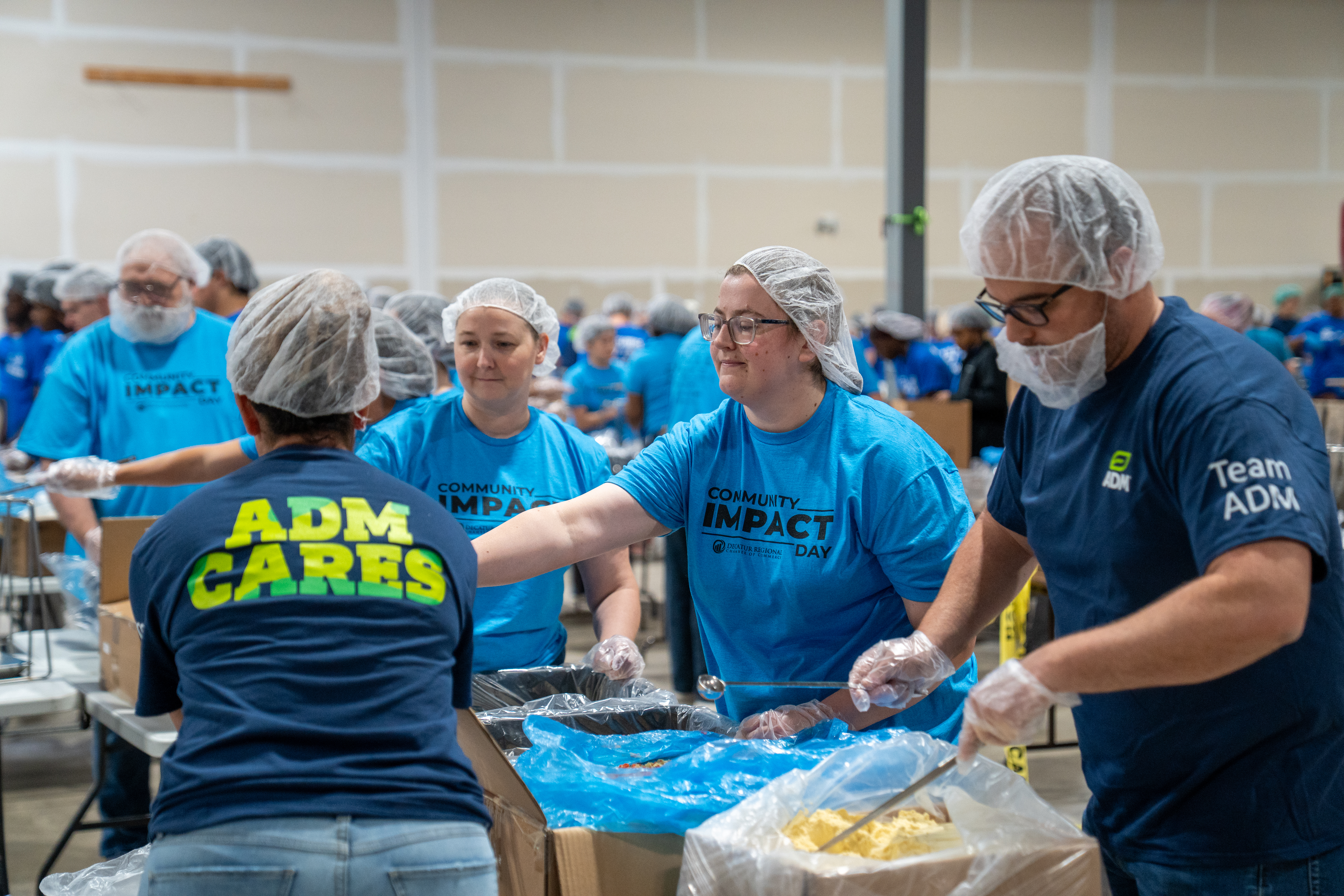 Volunteers wearing hairnets, gloves, and blue shirts work at food-packing stations inside a large warehouse. Several volunteers stand around a table measuring and packaging ingredients from large containers, while many others work in the background as part of an organized meal-packing effort. Visible shirt text includes “ADM CARES,” “Community Impact Day,” and “Team ADM.”