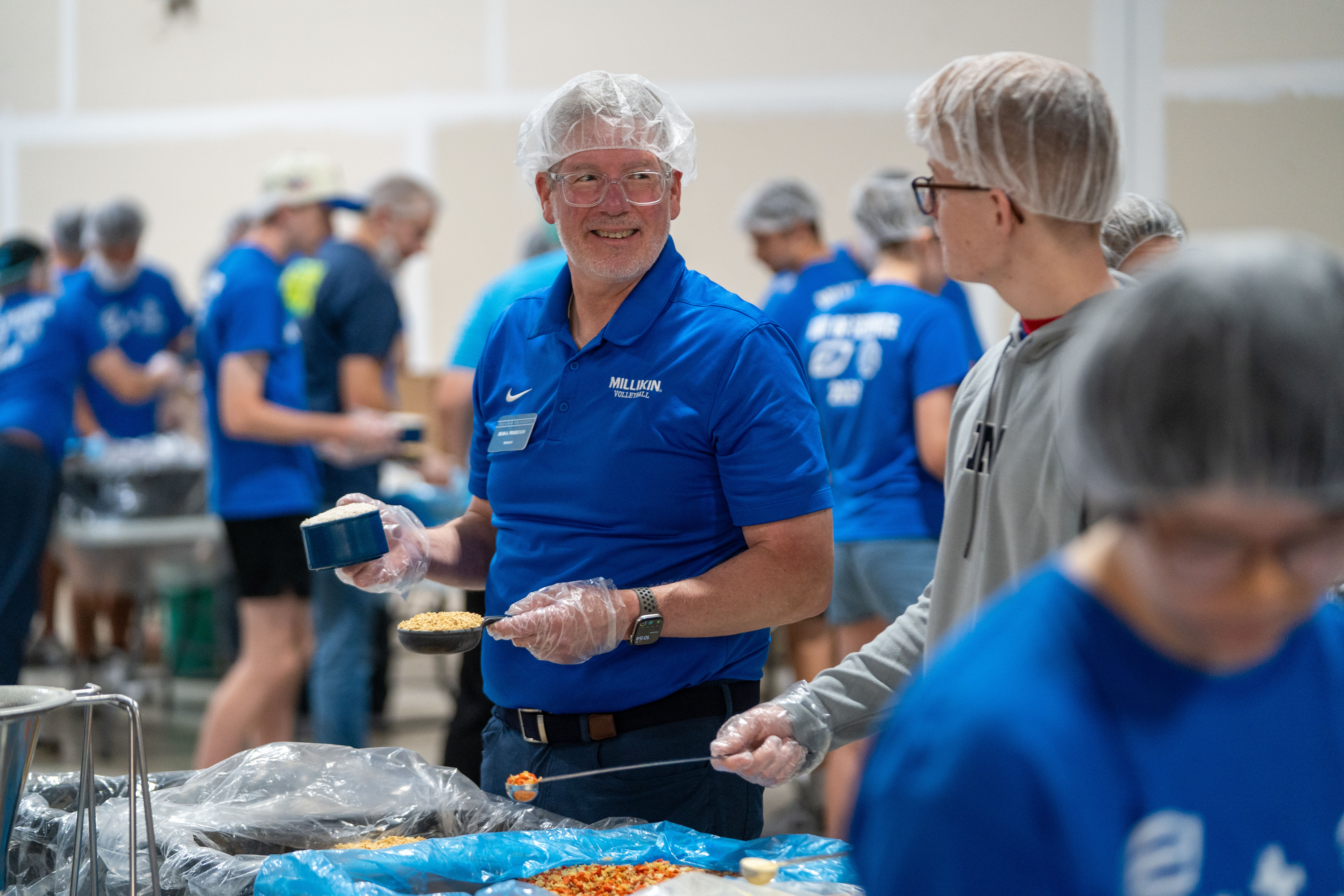 Volunteers wearing blue shirts, hairnets, and gloves participate in a meal-packing event inside a warehouse. In the foreground, one volunteer measures ingredients with scoops while standing beside large containers of dried food components. Additional volunteers work at assembly stations throughout the background.