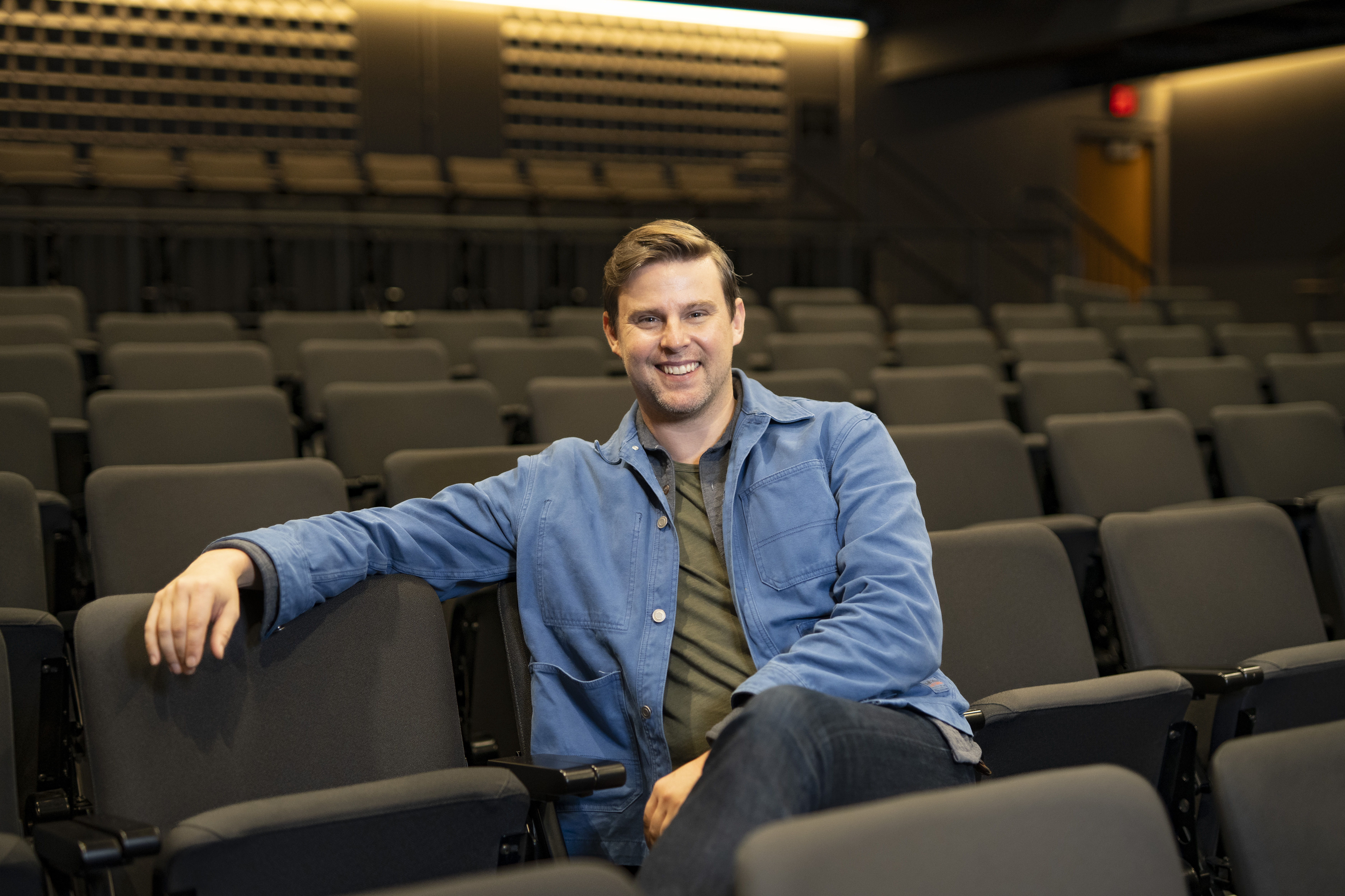 A person wearing a blue jacket, dark shirt, and jeans sits among rows of empty theatre seats, facing the camera. One arm rests across the back of a nearby chair while the other rests in their lap. The auditorium features dark seating, warm accent lighting, and tiered rows extending into the background. The shallow depth of field keeps the seated person in focus while the surrounding theatre interior appears softly blurred.