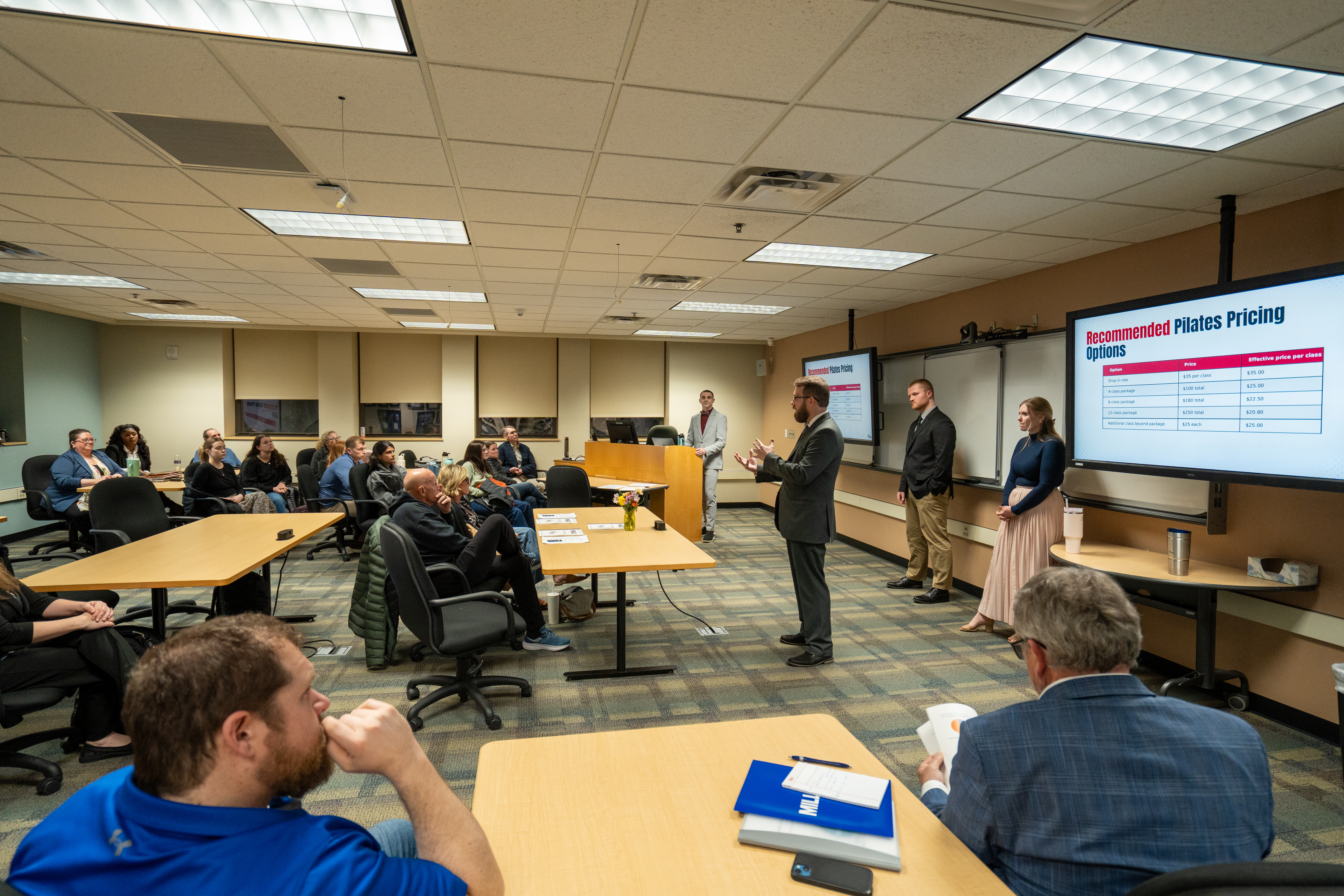 Conference room with attendees seated at tables while three presenters stand at the front of the room. Two large display screens show a presentation. A speaker in business attire addresses the audience, while another person stands at a podium nearby. The room has classroom-style seating and overhead fluorescent lighting.