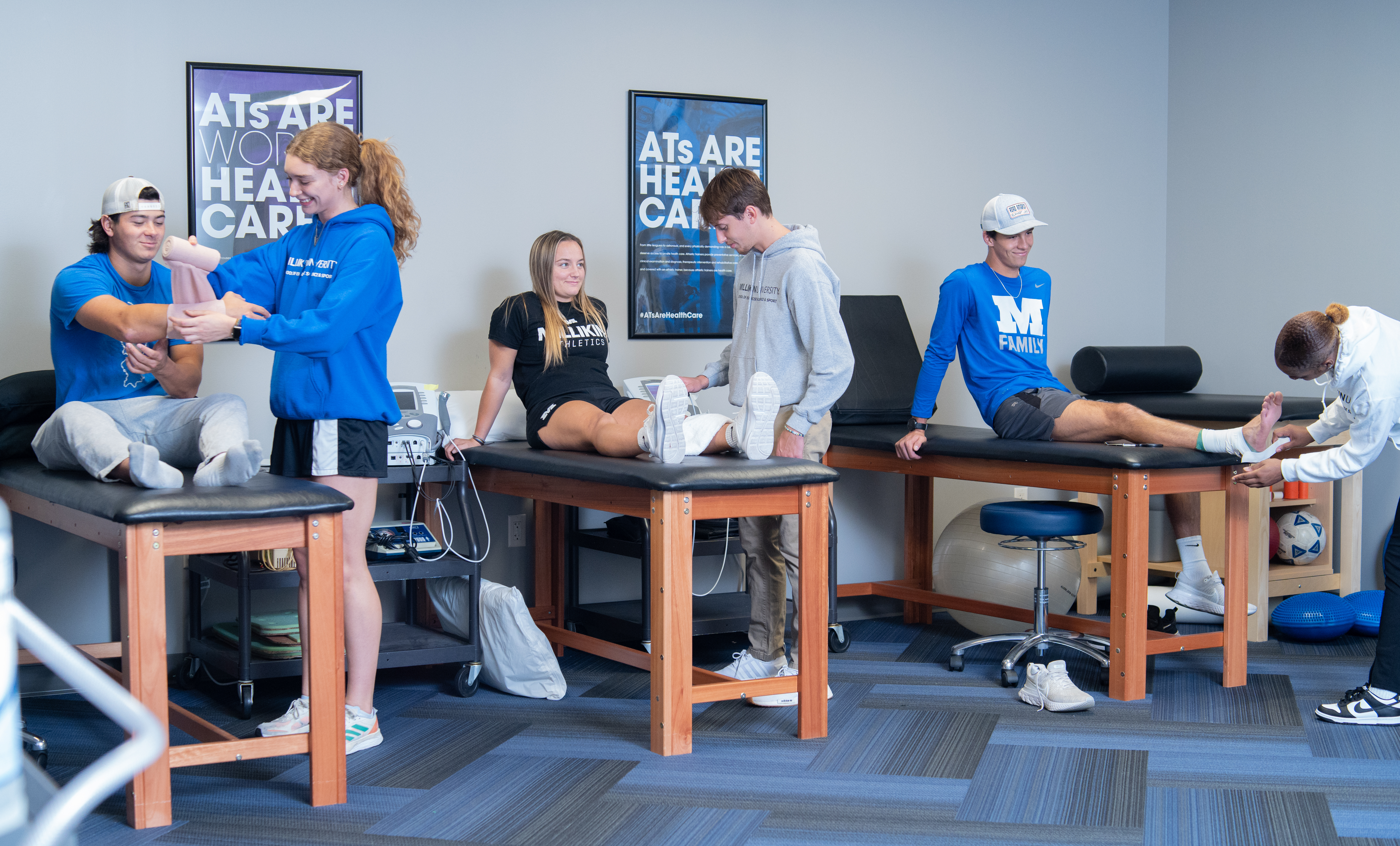 Several people participate in rehabilitation or athletic training activities in a clinical therapy room. Three treatment tables are arranged across the room, with individuals seated or lying on them while others assist with stretching, taping, or exercise instruction. Therapy equipment, stools, and exercise tools are visible around the tables. The room has blue-gray walls, blue patterned flooring, and posters related to health care on the wall.