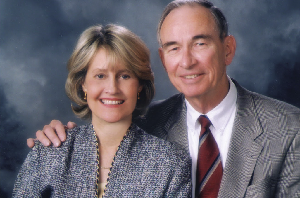Formal studio portrait of a woman and man posed side-by-side against a blue-gray backdrop. The woman wears a textured gray jacket and gold earrings, while the man wears a gray plaid suit jacket, white shirt, and striped tie, with a hand resting on the woman's shoulder. Both face the camera in a traditional professional portrait setting with soft, even lighting.