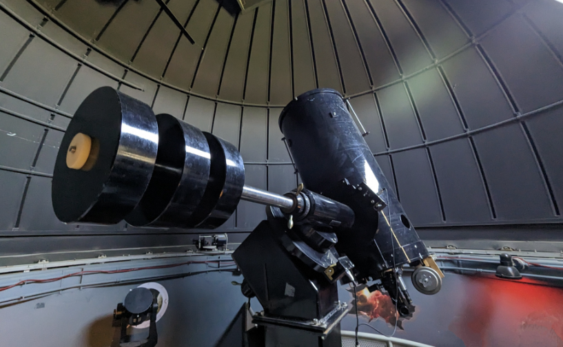 Large black telescope mounted inside a domed observatory. The telescope points upward beneath a curved metal dome with segmented panels, while observation equipment and control components are visible around the base. Soft interior lighting highlights the instrument's reflective surfaces and mechanical details.