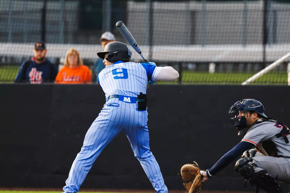 Baseball player wearing a blue-and-white pinstriped uniform with the number 9 stands in the batter’s box preparing to swing, while a catcher in gray protective gear crouches behind home plate. Spectators and a black outfield fence are visible in the background.