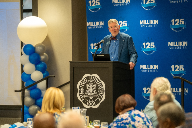 A speaker stands at a Millikin University podium addressing attendees at a banquet event, with a blue Millikin 125th anniversary backdrop and blue, white, and gray balloon display behind the stage.