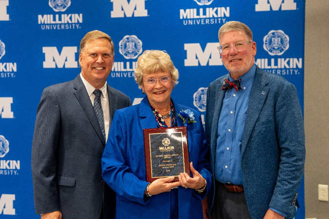Three individuals pose in front of a Millikin University backdrop, with the person in the center holding a commemorative plaque during an awards recognition ceremony.