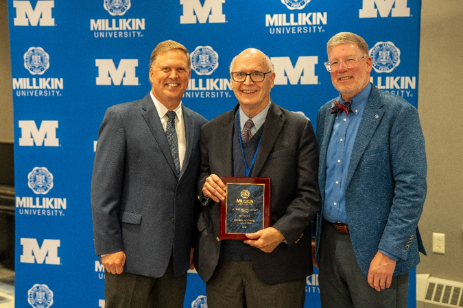Three individuals pose in front of a Millikin University backdrop, with the person in the center holding a recognition plaque during an awards presentation.