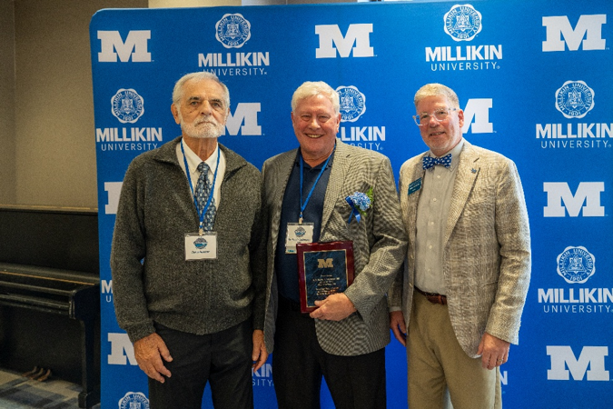 Three attendees pose for a photograph in front of a blue Millikin University backdrop featuring university logos and large “M” graphics. The person in the center holds a maroon-and-gold recognition plaque and wears a blue ribbon, while the two individuals standing on either side also wear event name badges. A piano is visible at the edge of the room, and the image appears to have been taken during a university awards or Hall of Fame recognition event.