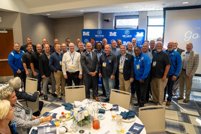 A large group of award recipients, alumni, and university representatives gather for a group photo during a Millikin University recognition event. The attendees stand in several rows in front of a blue Millikin University backdrop, many wearing event name badges and blue ribbons. In the foreground, banquet tables are set with plates, beverages, floral centerpieces, and event programs, capturing the atmosphere of a celebratory luncheon or awards ceremony.