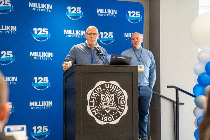 A speaker stands at a podium displaying the Millikin University seal while addressing attendees during a university recognition event. Another presenter stands nearby as the speaker delivers remarks. Behind them, a blue backdrop features Millikin University’s 125th anniversary branding, and a blue, white, and silver balloon column decorates the edge of the stage. The image captures a formal presentation taking place in a banquet and awards ceremony setting.