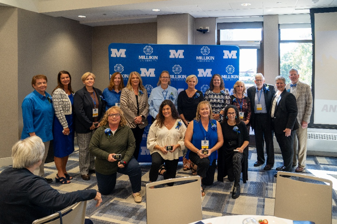 A group of award recipients and university representatives pose for a commemorative photo in front of a blue Millikin University backdrop. Four individuals kneel in the front row holding recognition plaques, while a larger group stands behind them wearing event badges and blue ribbons. The photo was taken during a banquet-style recognition program, with attendees seated at tables visible in the foreground and natural light streaming through large windows behind the group.