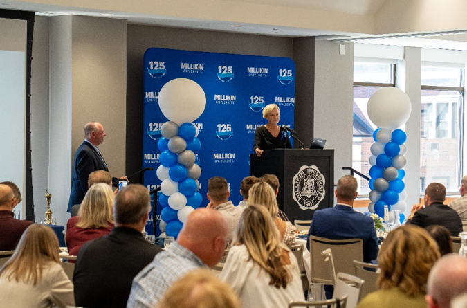 An attendee speaks from a podium bearing the Millikin University seal during a banquet-style recognition program. Guests seated at round tables face the stage, where the presenter stands between two blue, white, and silver balloon columns. A blue backdrop featuring Millikin University’s 125th anniversary branding frames the presentation area, while another attendee stands nearby and appears to be awaiting recognition. Large windows along the side of the room fill the event space with natural light.
