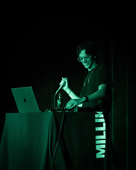 A performer stands behind a table with DJ and audio equipment on a dark stage, illuminated by green lighting. A laptop is open on the left side of the table, while the performer holds a microphone in one hand and adjusts controls on the mixer with the other. A water bottle sits near the equipment. The table covering displays MILLIKIN in large vertical white letters. Black stage curtains form the background, creating a simple concert or live performance setting.