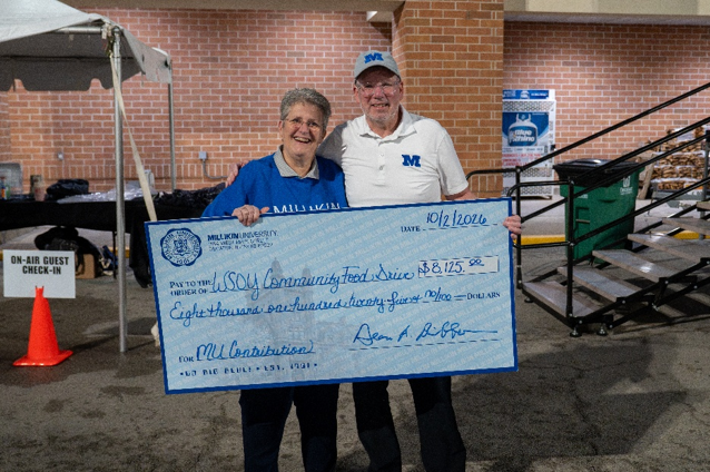 A woman in a blue sweatshirt and a man in a white polo shirt and hat with a blue M on it hold a large check. The check is written for $8,125 to the WSOY Community Food Drive from Millikin University.