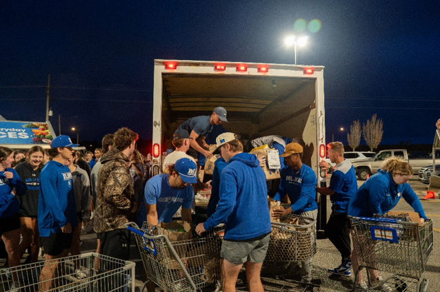 Several students dressed in blue unload boxes and bags of foods into waiting shopping carts.