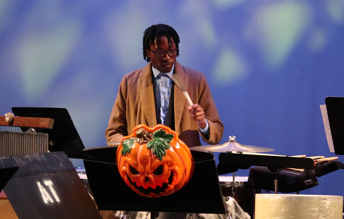 A student dressed as Dr. Who plays the drums back of stage with an evil pumpkin mask hanging off of a music stand in the foreground.