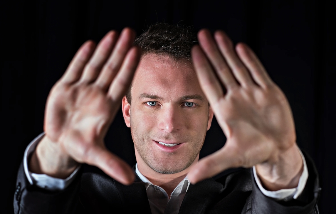 Close up of Jason Hudy, a white man, with a black background. His hands form a diamond in front of him, framing his face. 