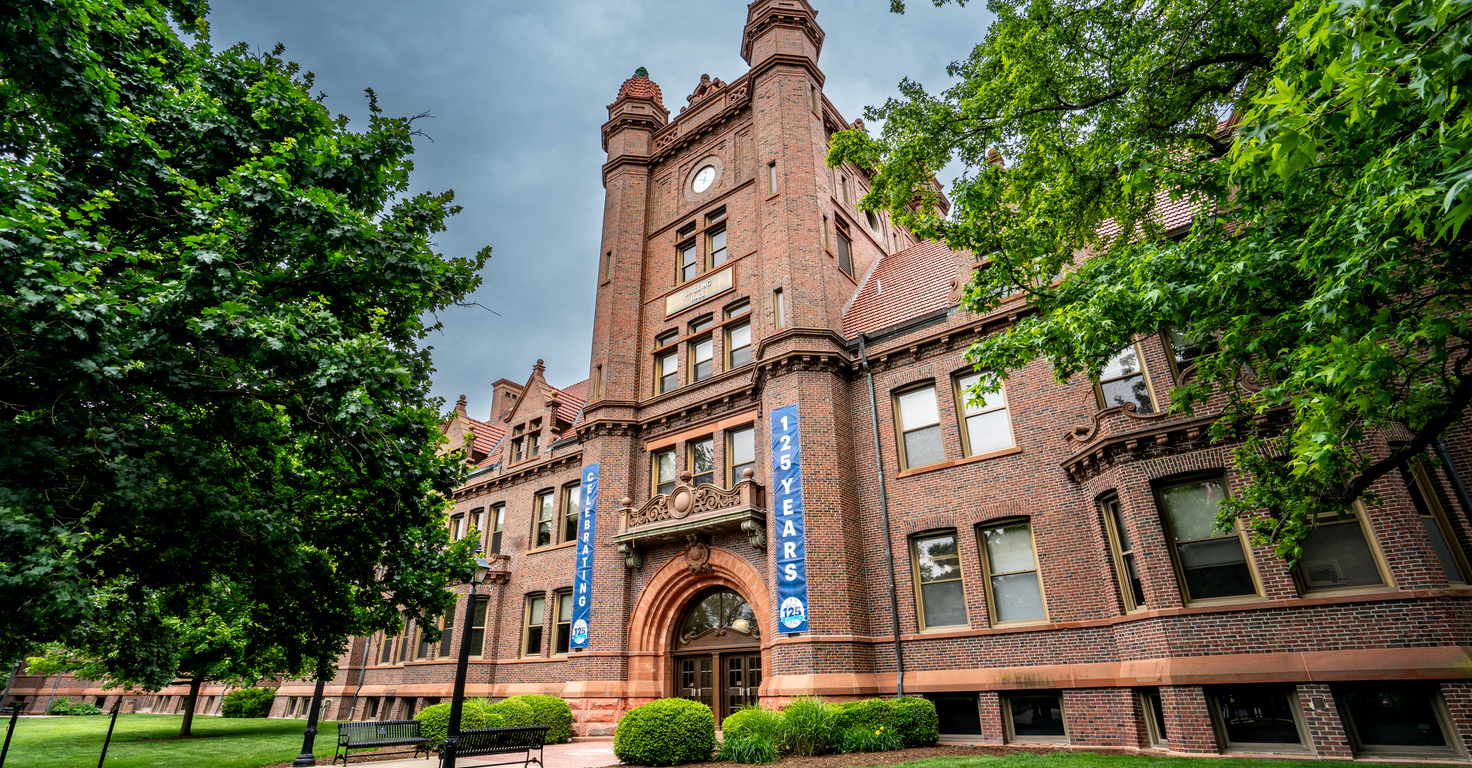 Historic brick building with ornate architectural details, clock tower, and arched entrance surrounded by green trees and lawn. Blue banners on the building celebrate its 125-year anniversary.