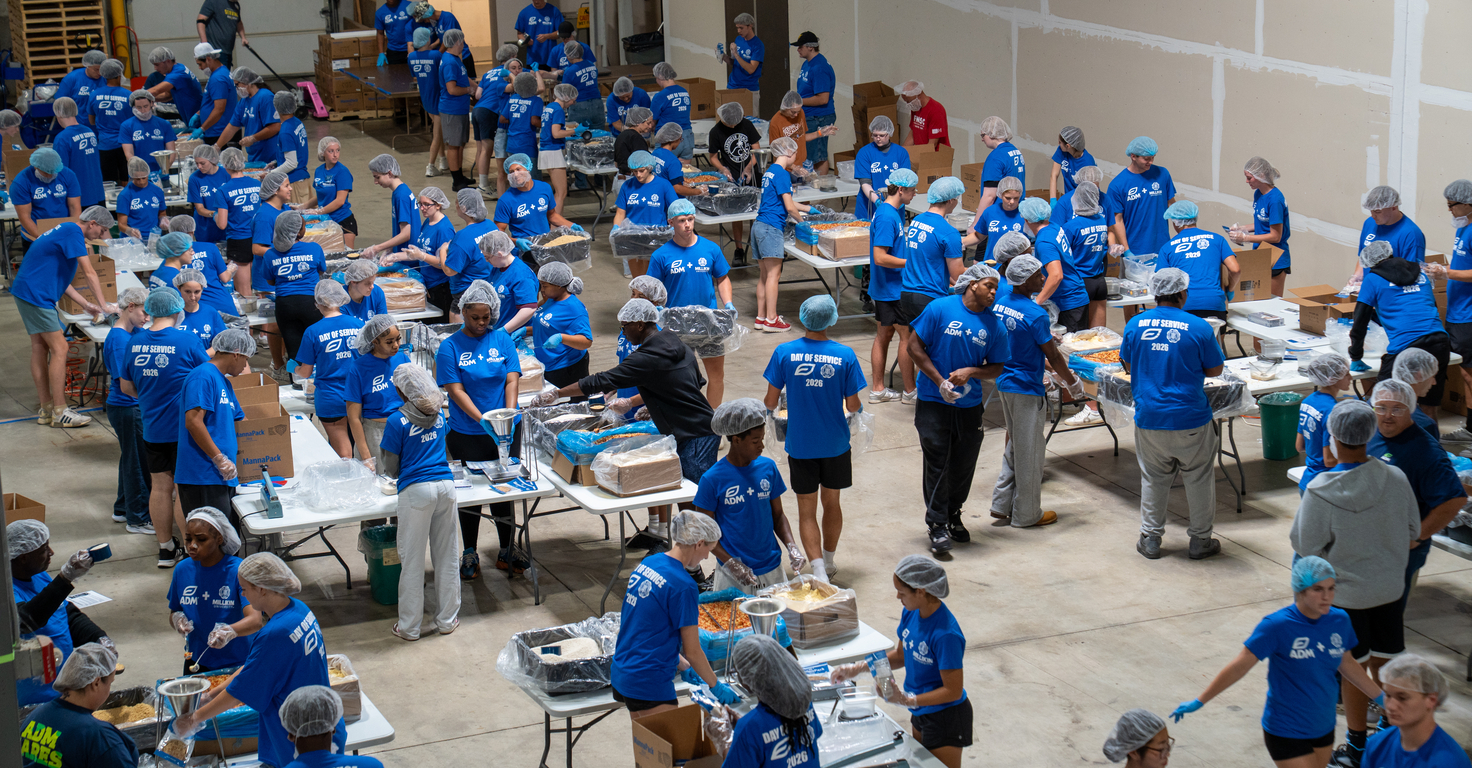 Large indoor warehouse space filled with dozens of volunteers wearing matching blue T-shirts and hairnets, working at long tables to assemble and package meals. Boxes, trays, and food-packaging materials are spread across the tables, while volunteers move between stations in an organized assembly-line setup. The high-ceiling room has unfinished walls, industrial lighting, and stacks of supplies visible in the background.