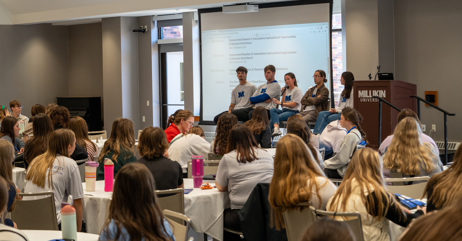 Audience members seated at round tables listen to a panel discussion at a conference room. Five panelists sit at the front near a podium labeled “Millikin University,” with a presentation agenda projected on a screen behind them. Attendees face the speakers, and several water bottles and notebooks are visible on the tables throughout the room.