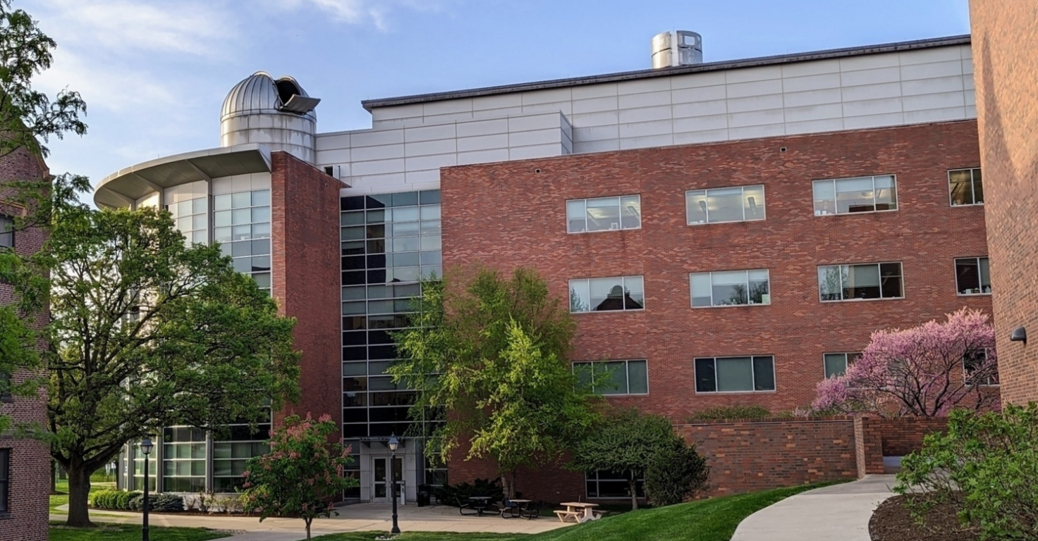 Modern multi-story red brick academic or office building with large glass windows and a central glass entrance. Mature green trees and landscaping frame the building, with a curved sidewalk leading through the foreground under a clear blue sky.