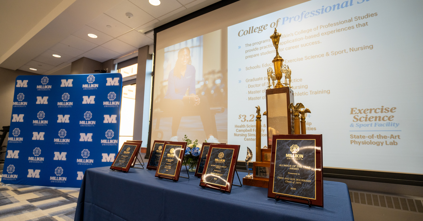 Awards plaques and a championship trophy displayed on a blue-draped table in a conference room, with a Millikin University backdrop and presentation screen in the background.
