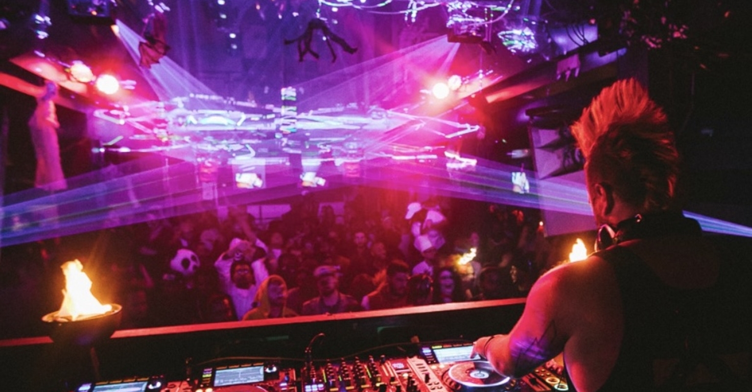 A DJ performs from an elevated booth overlooking a crowded nightclub illuminated by vivid pink, purple, and red lighting. Multiple laser beams stretch across the room above the audience, while professional DJ equipment with glowing controls fills the foreground. Small flame effects burn on either side of the booth, and the dark venue ceiling is lined with lighting rigs and suspended decorations. The scene captures a high-energy live music event with dramatic lighting and a packed dance floor.