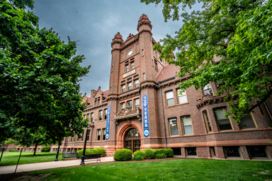 Historic brick building with ornate architectural details, clock tower, and arched entrance surrounded by green trees and lawn. Blue banners on the building celebrate its 125-year anniversary.