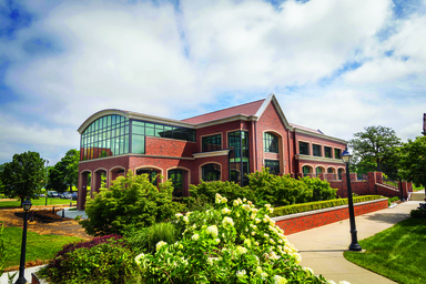 Brick academic building with large glass windows, landscaped shrubs, and white flowers in the foreground, under a partly cloudy sky.