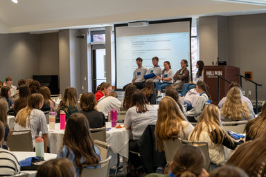 Audience members seated at round tables listen to a panel discussion at a conference room. Five panelists sit at the front near a podium labeled “Millikin University,” with a presentation agenda projected on a screen behind them. Attendees face the speakers, and several water bottles and notebooks are visible on the tables throughout the room.