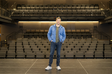 A person stands at the center of a theatre stage facing the camera, wearing a blue jacket, dark jeans, and light-colored shoes. Rows of empty auditorium seats fill the foreground and middle ground, while tiered seating and balconies rise in the background. Warm overhead lighting illuminates the modern performance venue, highlighting the symmetry of the stage and audience seating.