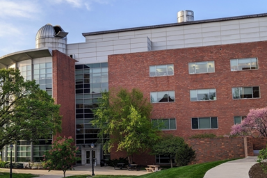 Modern multi-story red brick academic or office building with large glass windows and a central glass entrance. Mature green trees and landscaping frame the building, with a curved sidewalk leading through the foreground under a clear blue sky.