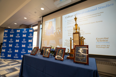 Awards plaques and a championship trophy displayed on a blue-draped table in a conference room, with a Millikin University backdrop and presentation screen in the background.