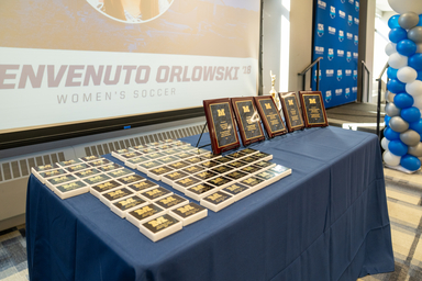 A display table covered with a blue tablecloth holds dozens of black-and-gold awards and five larger framed plaques arranged in rows. Behind the table, a large projection screen displays “Benvenuto Orlowski ’18 Women’s Soccer.” A blue-and-white balloon column and a branded backdrop stand to the right, suggesting an athletic awards or Hall of Fame recognition event.