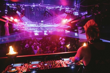 A DJ performs from an elevated booth overlooking a crowded nightclub illuminated by vivid pink, purple, and red lighting. Multiple laser beams stretch across the room above the audience, while professional DJ equipment with glowing controls fills the foreground. Small flame effects burn on either side of the booth, and the dark venue ceiling is lined with lighting rigs and suspended decorations. The scene captures a high-energy live music event with dramatic lighting and a packed dance floor.
