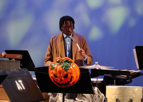 A student dressed as Dr. Who plays the drums back of stage with an evil pumpkin mask hanging off of a music stand in the foreground.