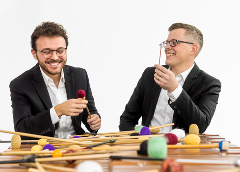 The two male members of pax duo hold mallets and smile.