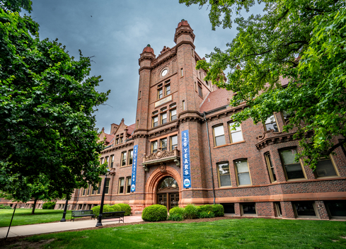Historic brick building with ornate architectural details, clock tower, and arched entrance surrounded by green trees and lawn. Blue banners on the building celebrate its 125-year anniversary.
