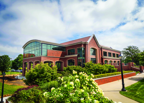 Brick academic building with large glass windows, landscaped shrubs, and white flowers in the foreground, under a partly cloudy sky.