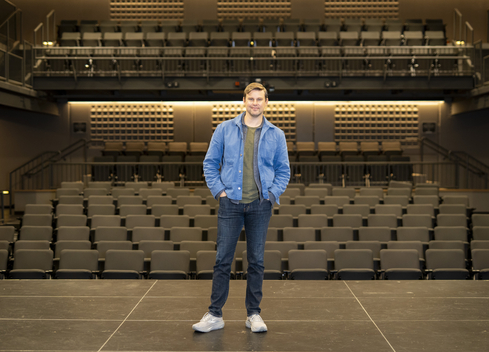 A person stands at the center of a theatre stage facing the camera, wearing a blue jacket, dark jeans, and light-colored shoes. Rows of empty auditorium seats fill the foreground and middle ground, while tiered seating and balconies rise in the background. Warm overhead lighting illuminates the modern performance venue, highlighting the symmetry of the stage and audience seating.