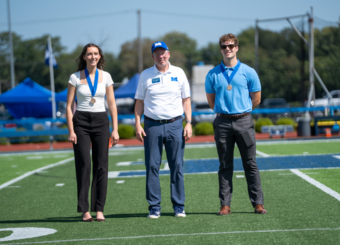 Three individuals wearing medals stand on a football field during an outdoor ceremony. The person in the center wears a white polo shirt and blue cap with an “M” logo, while the two individuals on either side wear business-casual attire. Blue tents, field equipment, and trees are visible in the background under a clear sky.