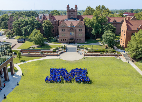 Aerial view of a college campus courtyard on a sunny day. Hundreds of students wearing blue shirts stand on a large green lawn arranged in the shape of a capital “M,” with historic red-brick academic buildings, trees, walkways, and landscaped terraces in the background.