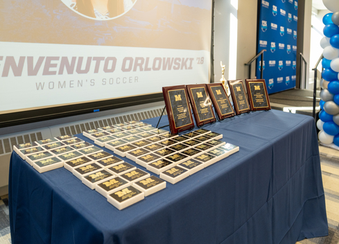 A display table covered with a blue tablecloth holds dozens of black-and-gold awards and five larger framed plaques arranged in rows. Behind the table, a large projection screen displays “Benvenuto Orlowski ’18 Women’s Soccer.” A blue-and-white balloon column and a branded backdrop stand to the right, suggesting an athletic awards or Hall of Fame recognition event.