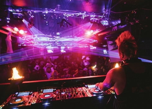 A DJ performs from an elevated booth overlooking a crowded nightclub illuminated by vivid pink, purple, and red lighting. Multiple laser beams stretch across the room above the audience, while professional DJ equipment with glowing controls fills the foreground. Small flame effects burn on either side of the booth, and the dark venue ceiling is lined with lighting rigs and suspended decorations. The scene captures a high-energy live music event with dramatic lighting and a packed dance floor.