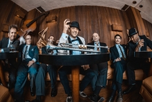 Band Members of Cherry Poppin Daddies sit at tables in a wood paneled room. A trumpet sits on the table in the middle. 