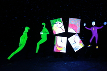 Two people in neon green body suits are illuminated by black light next to four neon paintings and a third figure with a purple bodysuit and white mask. 