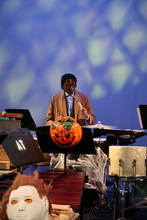 A student dressed as Dr. Who plays the drums back of stage with an evil pumpkin mask hanging off of a music stand in the foreground.